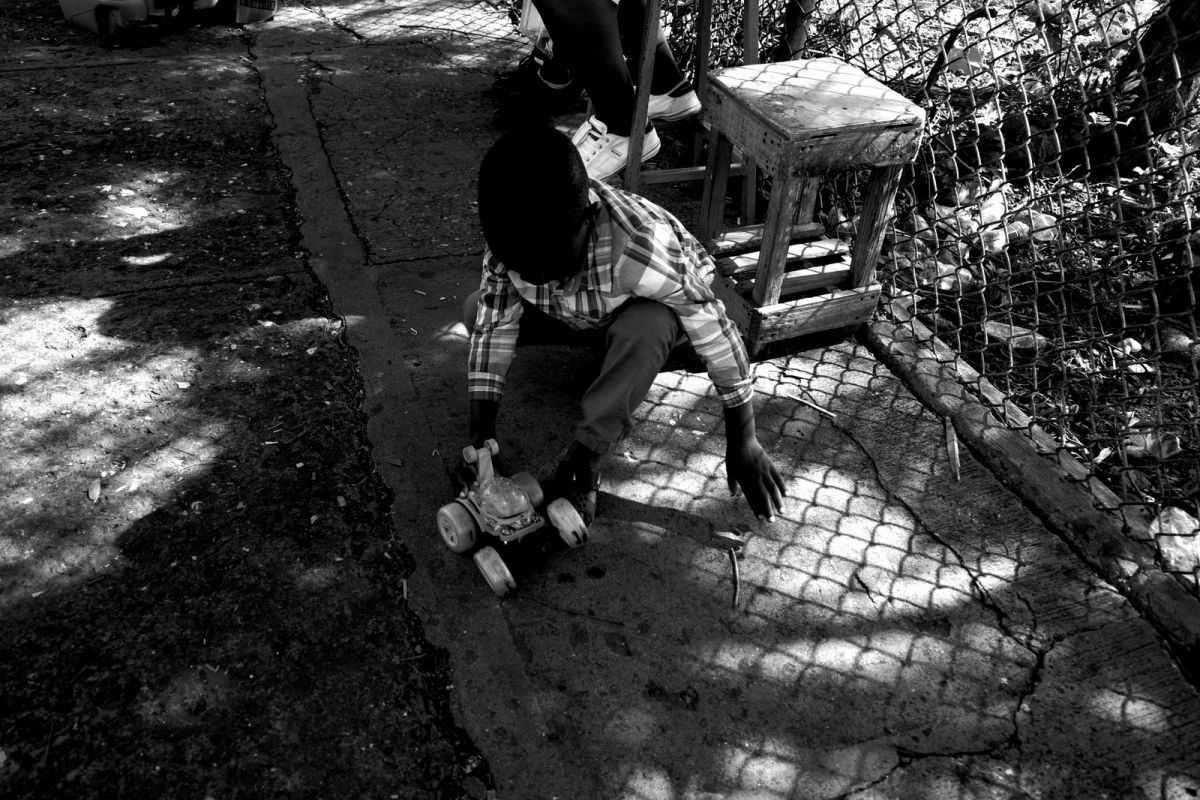 Child playing with his toy car in what looks like his Sunday best.  Being in the middle of a busy thoroughfare he probably wishes he were home. 